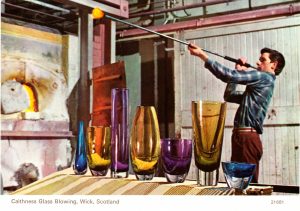 postcard showing glass blower in background with a row of vases in the foreground