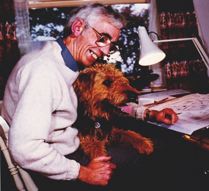 A man wearing glasses and smiling sitting at his desk with a brown terrier type dog on his lap on his desk there is a desk lamp and a collection of cartoon illustrations he is working on