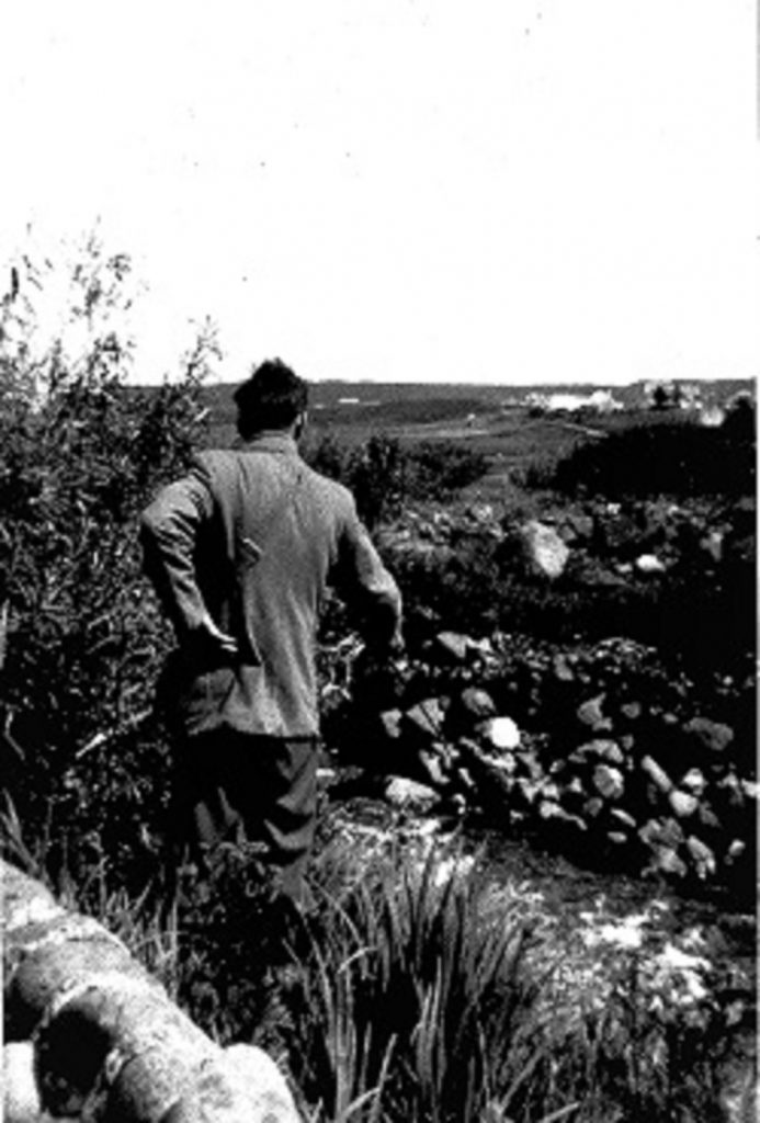 A man with his back to the camera fishing in the river or stream that runs in front of him he has his left hand resting on his hip and a fishing rod or net in his right hand