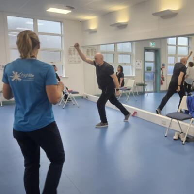 Instructor leading an exercise session in a bright fitness studio with participants performing standing movements.