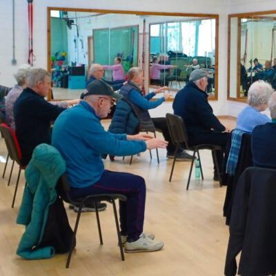 Group of older adults seated in a fitness studio following an instructor leading arm exercises in front of mirrors.