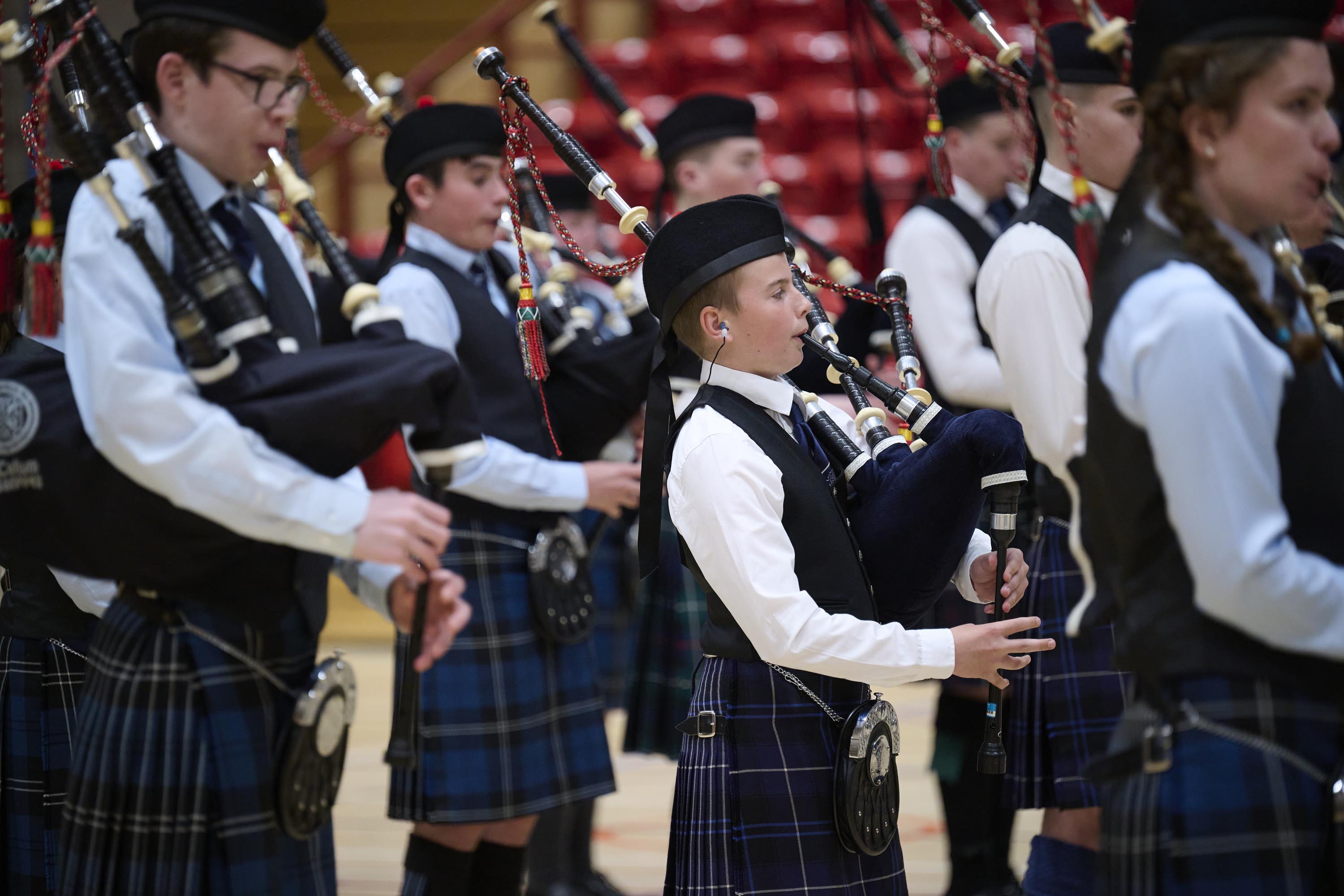 A group of young people playing the bagpipes and marching in a gym hall setting. They are wearing dark blue kilts, pale shirts and black waistcoats.