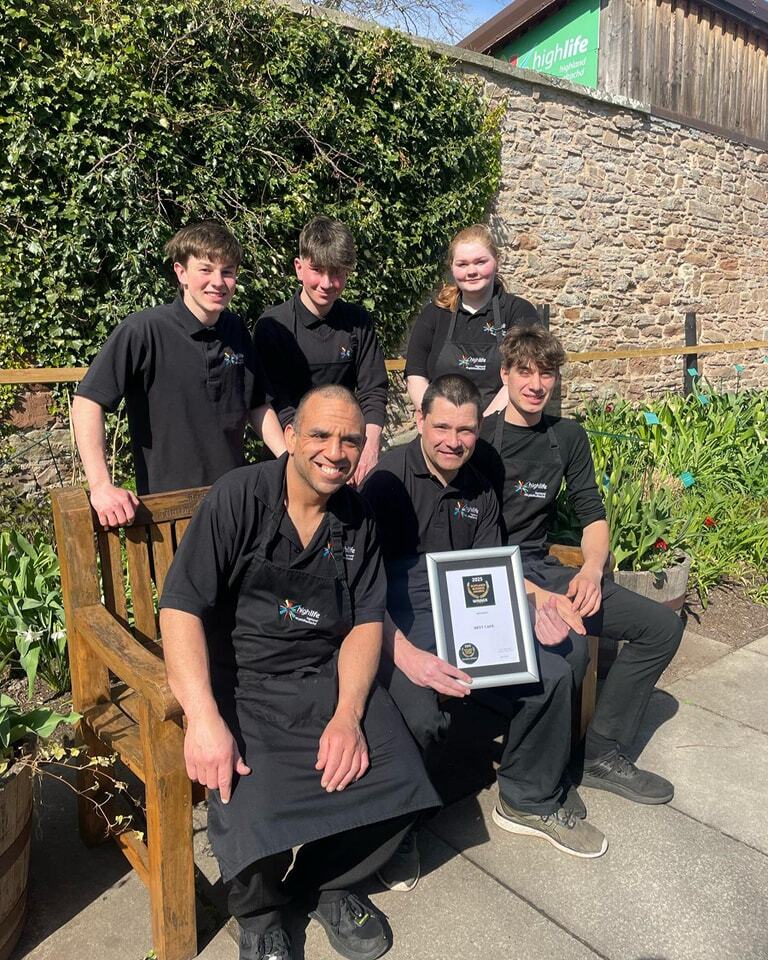 A group of 6 café staff sat in the sun showing off their framed award.