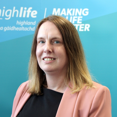 A woman smiles at the camera. She has brown hair and is wearing a pink suit