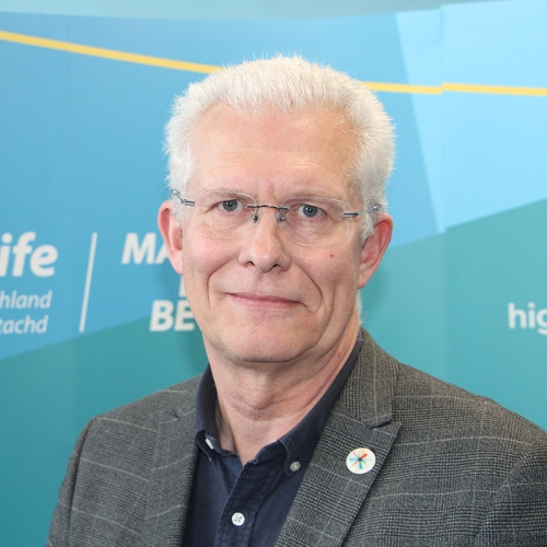 A man with grey hair looks at the camera. He is wearing a tweed suit with a blue shirt. He is wearing glasses.