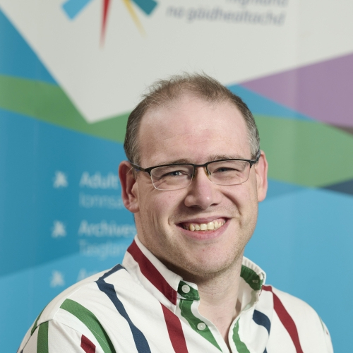 A man smiles at the camera. He is wearing a white shirt with large blue, green and red stripes.