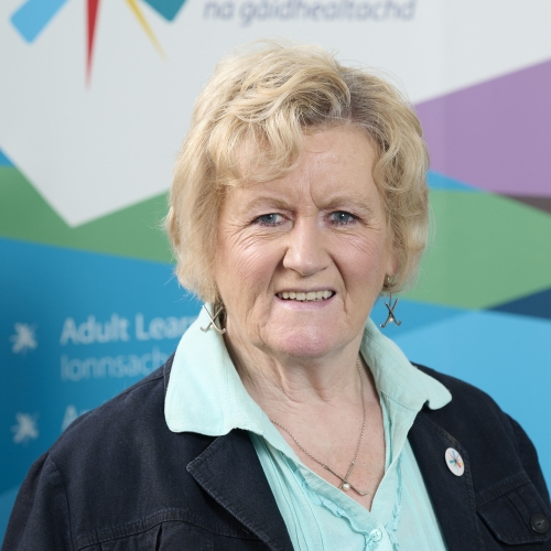 A woman smiles at the camera. She has short curled hair and is wearing a black jacket and minty-blue shirt.