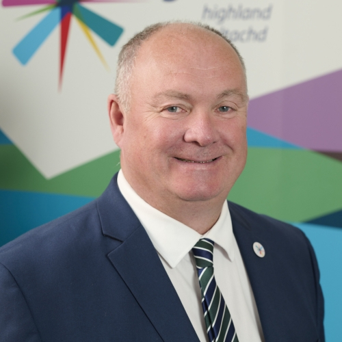 A man smiles at the camera. He is wearing a blue suit jacket, white shirt and striped tie.