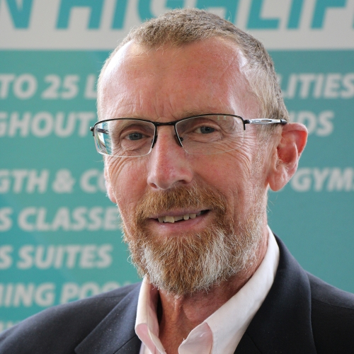 A man smiles at the camera. He is wearing glasses and a black suit.