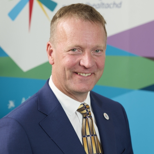 A white man with blonde hair smiles at the camera. He is photographed wearing a blue suit with a white pin on the lapel. He is wearing a yellow patterned tie.