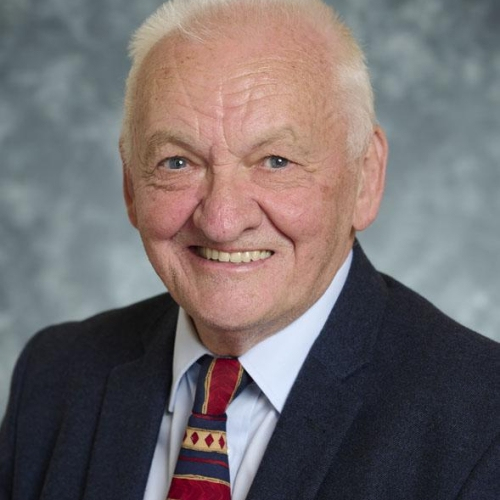 A man smiles at the camera. He has white hair and is wearing a black suit and a patterned blue, yellow and red tie.