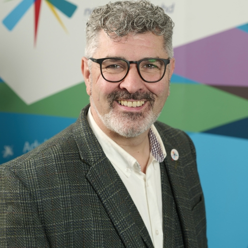 A man with grey curly hair smiles at the camera. He is wearing glasses, a checkered green suit jacket and a white shirt. He is wearing a High Life Highland badge on his lapel.