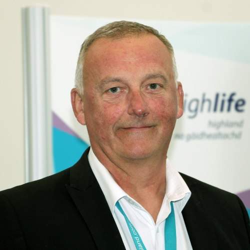 A white man smiles at the camera. He is wearing a black suit jacket, a white shirt and has a blue lanyard around his neck.
