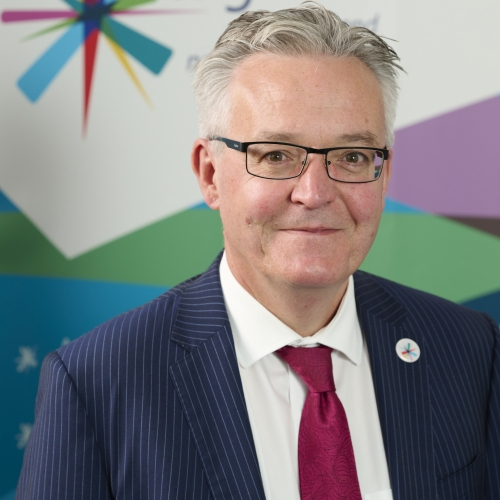 A man with grey smiles at the camera. He is wearing a blue pinstripe suit with a white shirt, red tie and glasses.