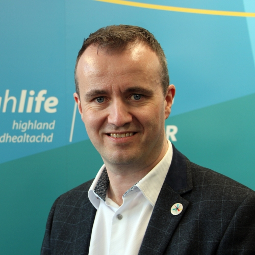 A man smiles at the camera. He is wearing a black suit and white shirt.