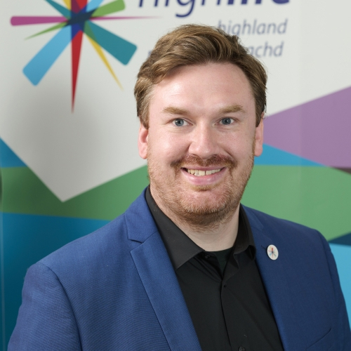 A man in the blue suit smiles at the camera. He has brown hair, a moustache and a beard.