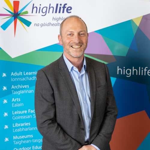 A man smiles at the camera. He is wearing a grey suit and blue shirt.