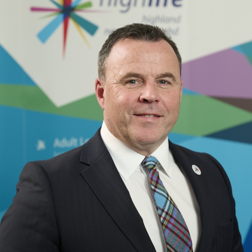 A white man with brown hair smiles at the camera. He is shown shoulders up and wearing a black suit jacket, a white shirt and a colourful tie.