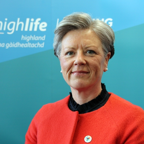 A woman smile sat the camera. She has short grey hair and is wearing a striking red suit.