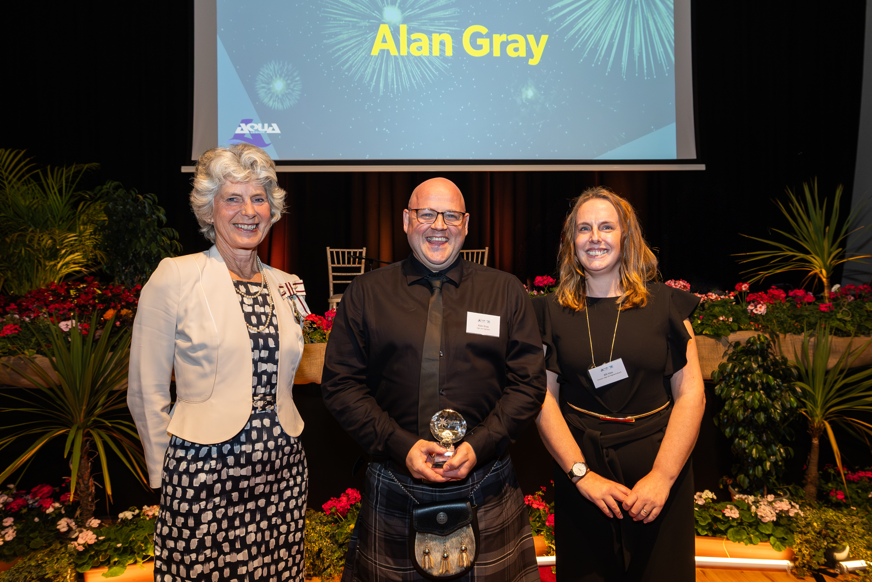 Three people standing on a stage at an awards event, with one holding a glass award and a presentation screen showing a name behind them.
