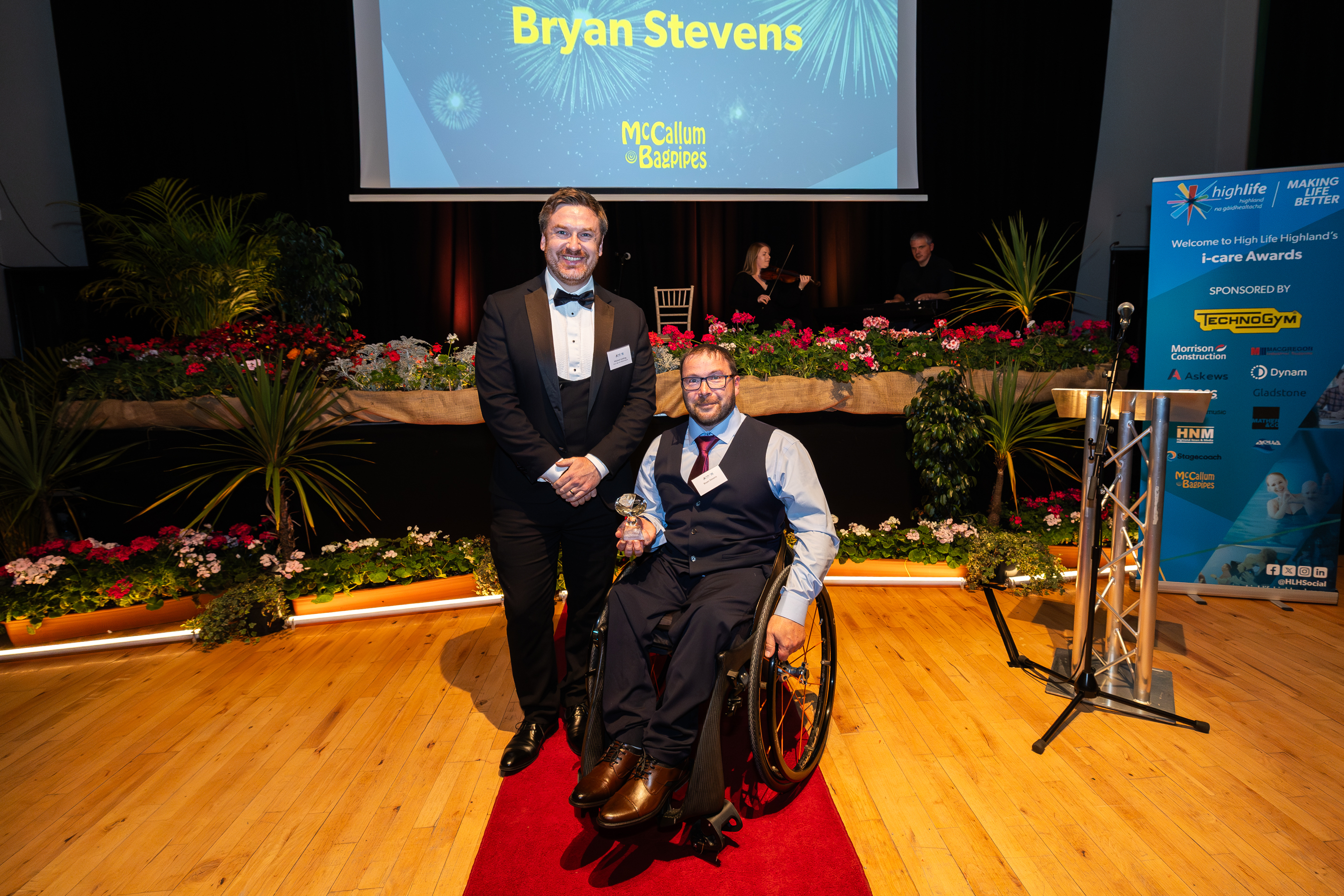 Two people posing on a red carpet at an awards event, one seated in a wheelchair holding a glass award, with a presentation screen behind them