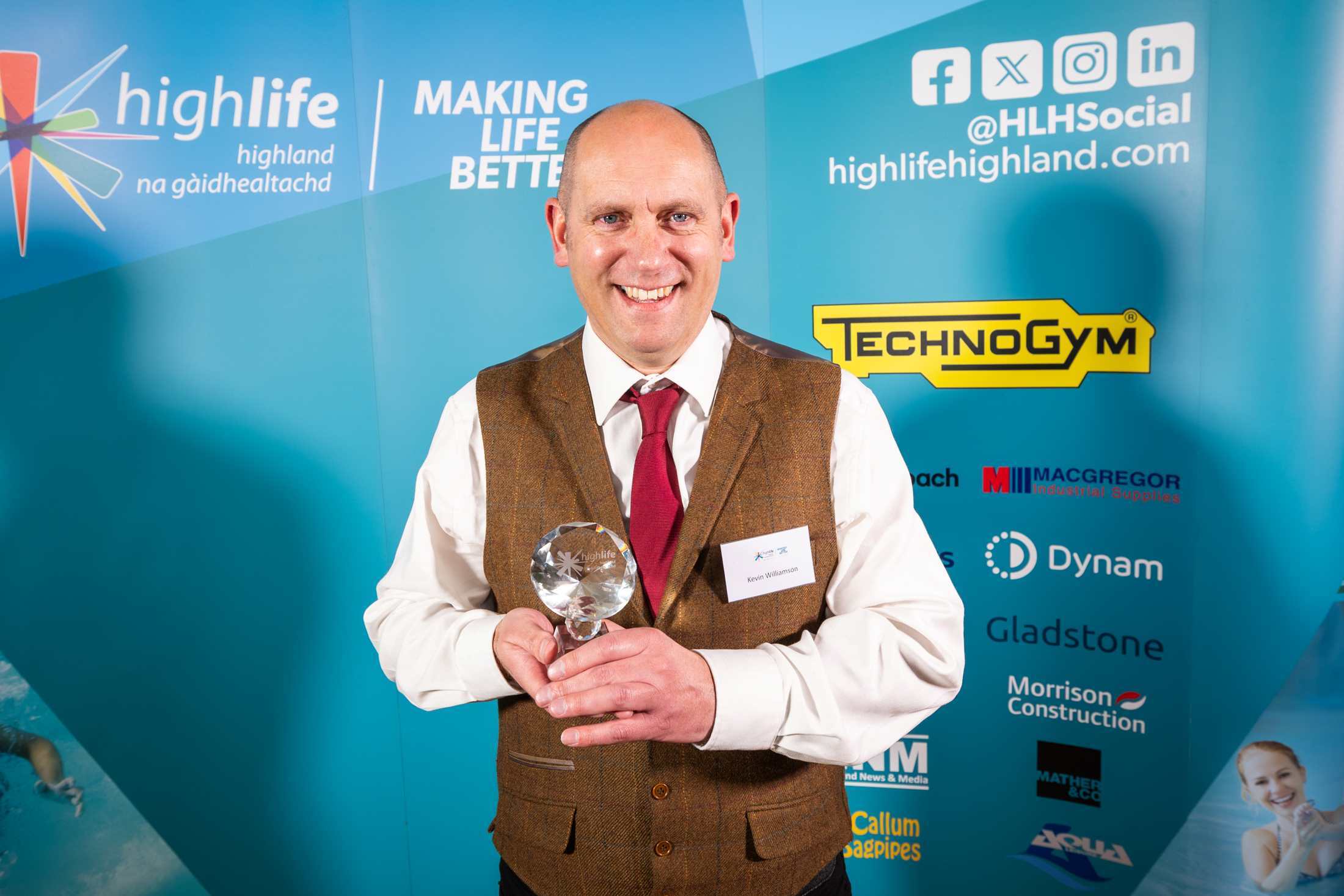 A person holding a glass award while standing in front of a High Life Highland event backdrop with sponsor logos.