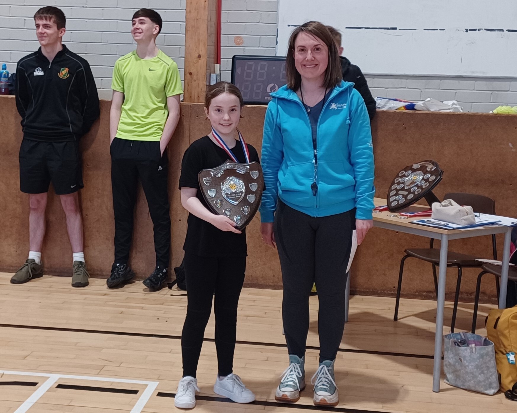 A young school pupil holding a winners shield. She is wearing dark clothing and standing beside and adult female in black leggings and a blue jacket. They are in a school hall setting and there are two older boys in the background.