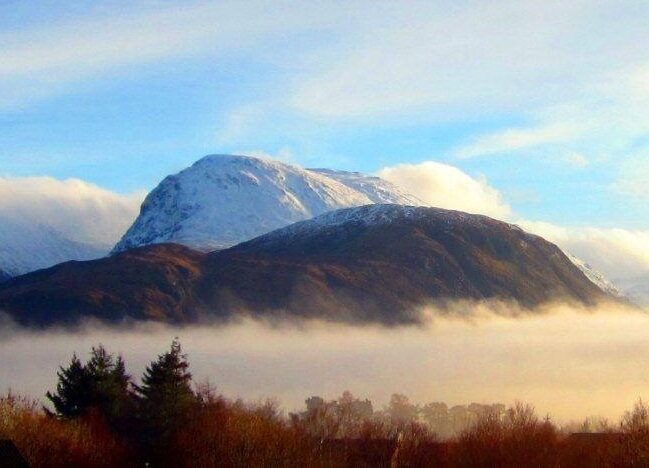 Snow-covered Ben Nevis with surrounding hills, blue sky, and mist over the valley.