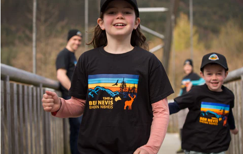 Children wearing matching Ben Nevis t‑shirts running across a wooden footbridge, with an adult walking behind them.
