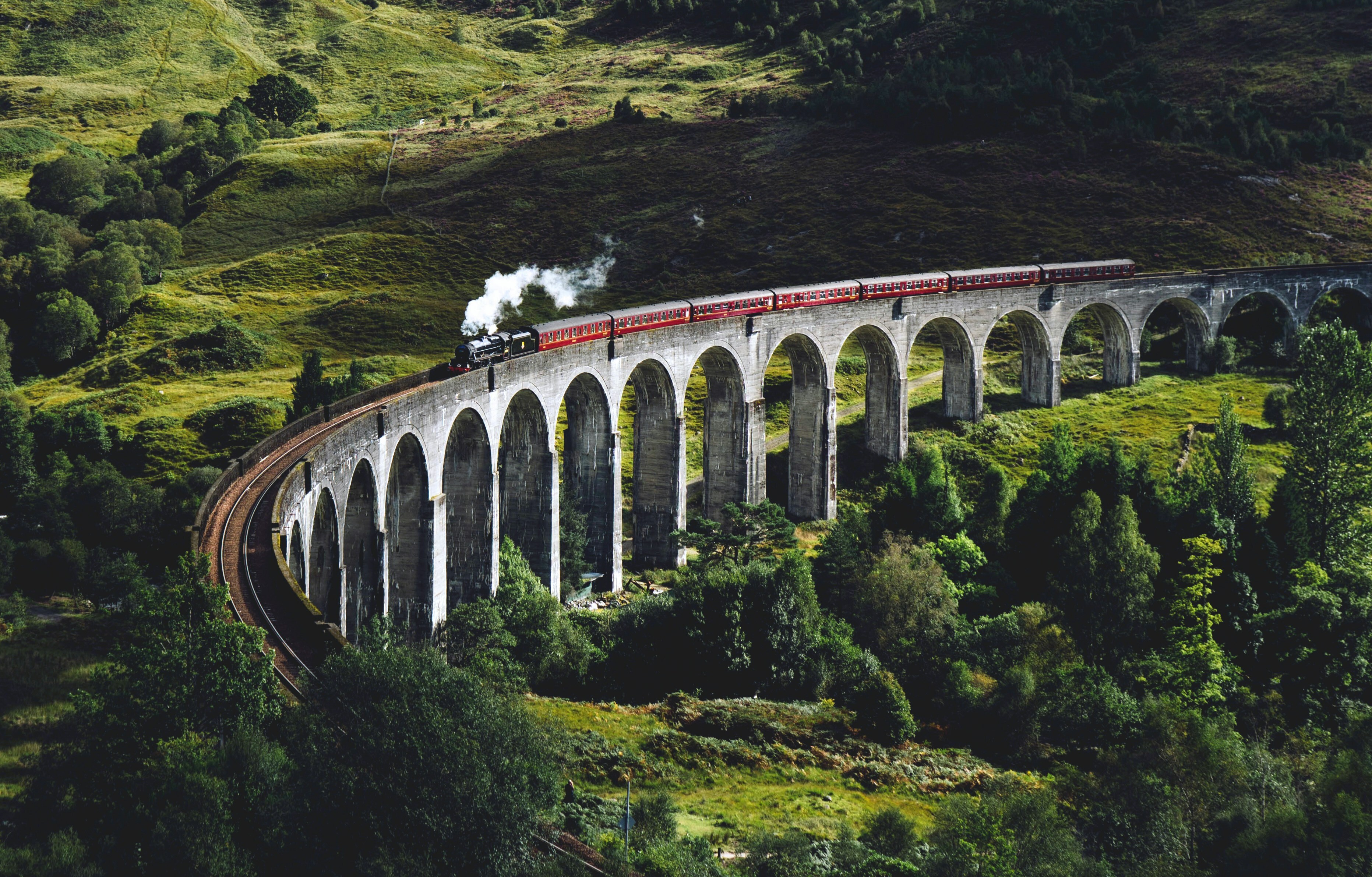 A steam train crosses a long stone viaduct surrounded by green hills.