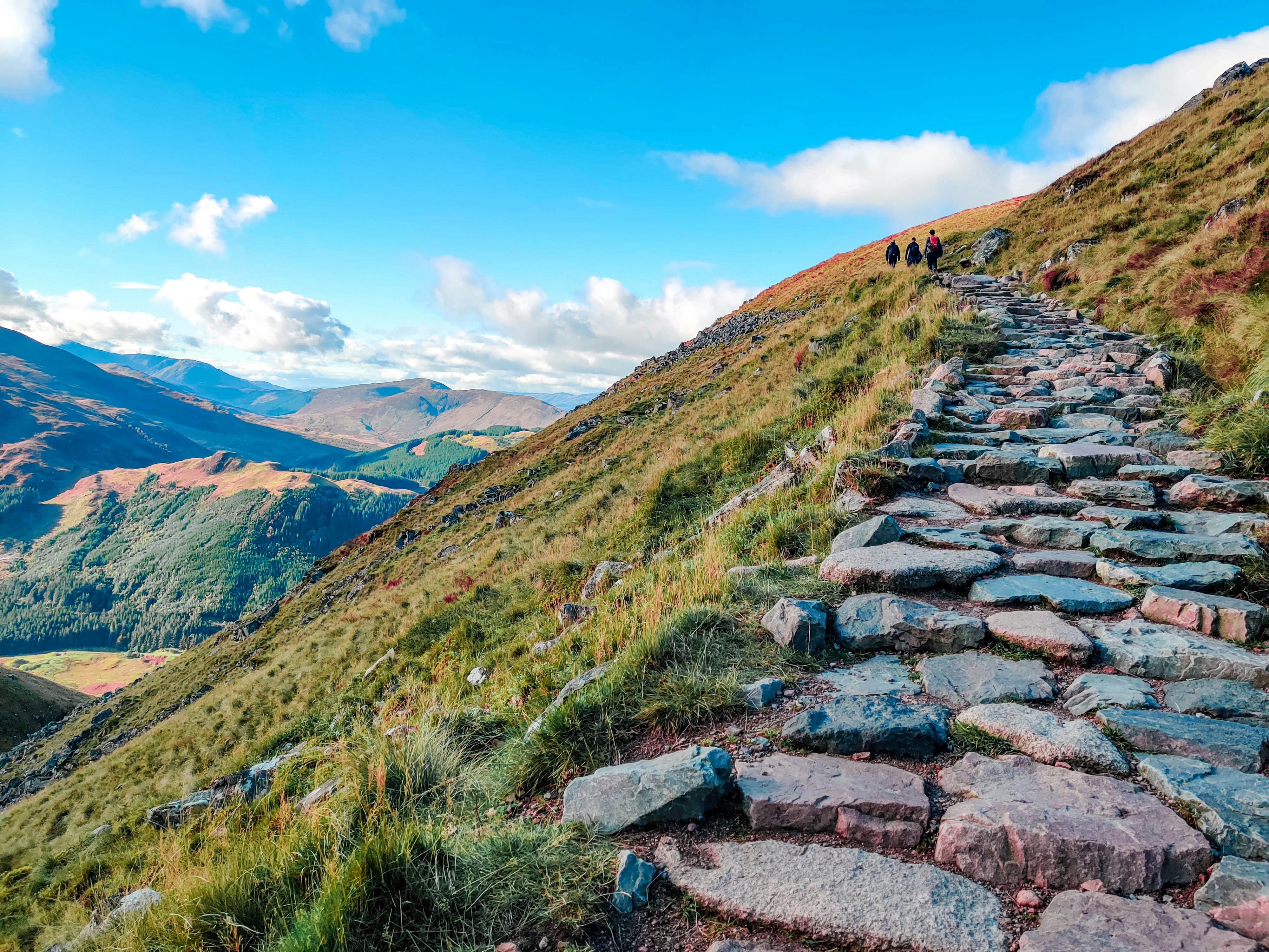 A rocky mountain path climbs uphill with a few hikers in the distance.
