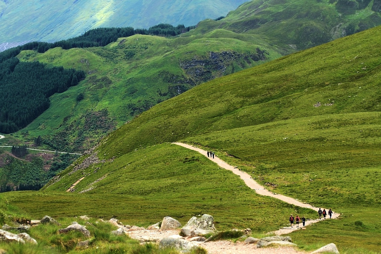 A winding path stretches across grassy hills with small groups of hikers walking along it.