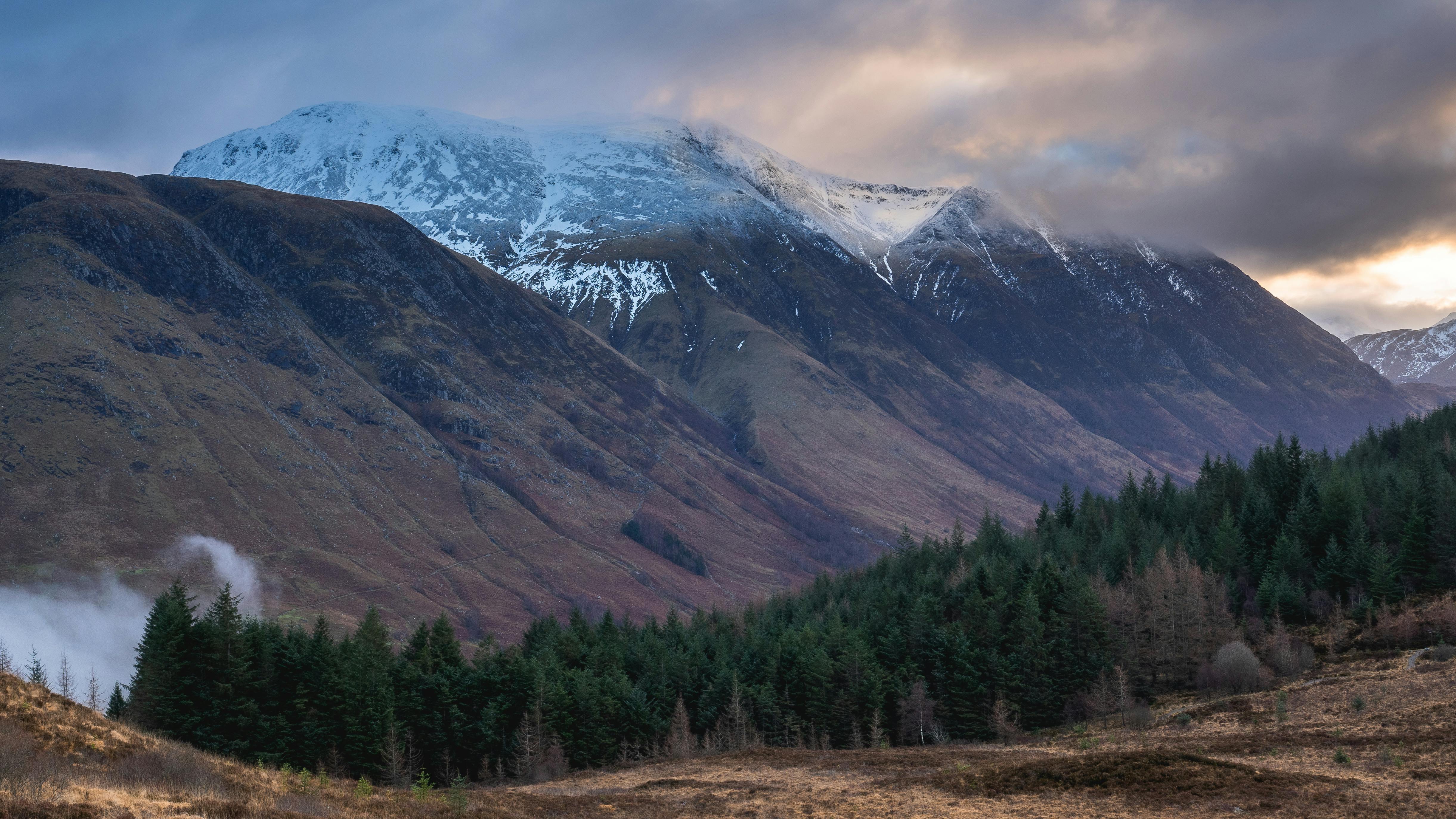 Snow‑topped mountains rise above dark green forest at the base, under a cloudy sky.