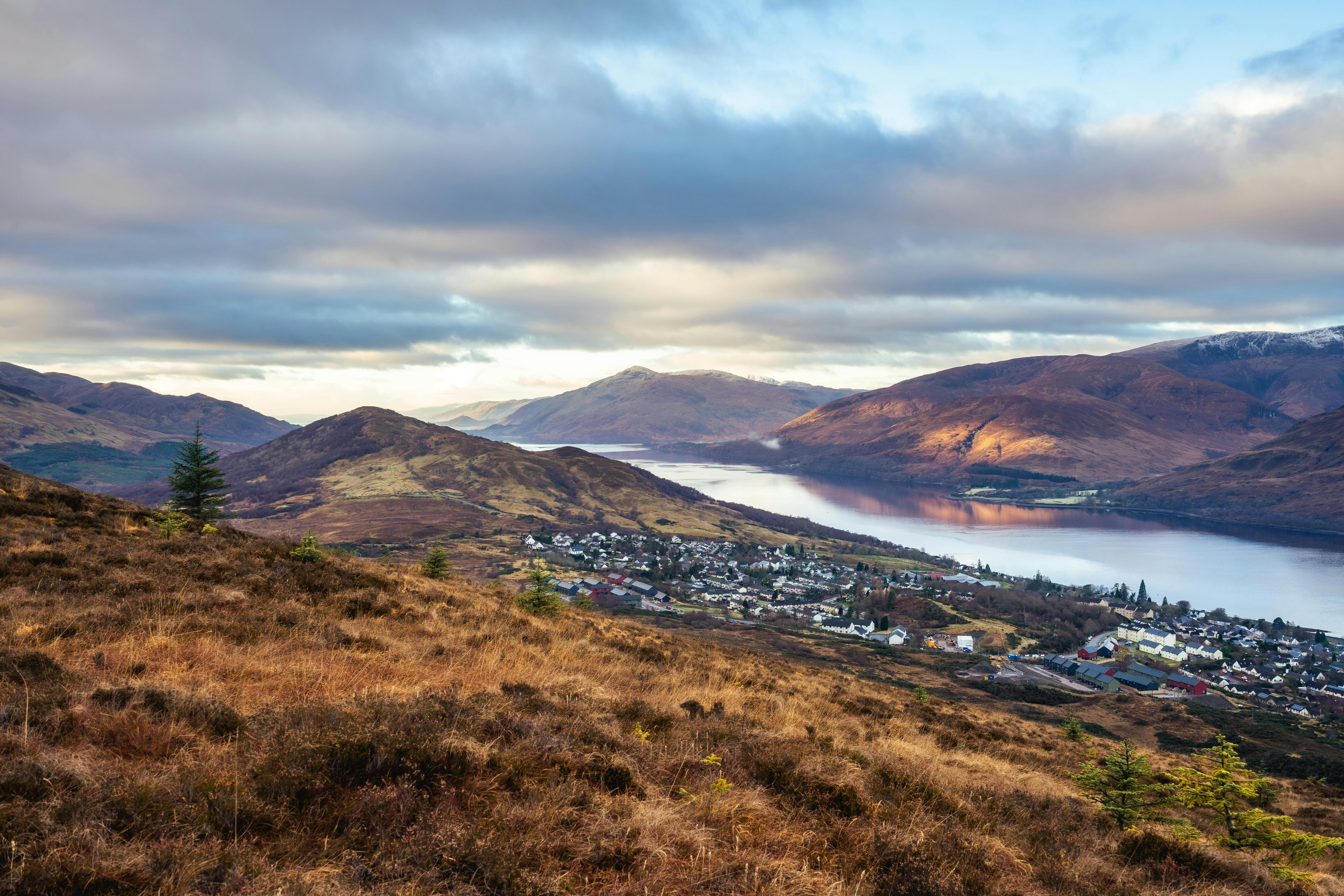 A loch and small town sit in a valley surrounded by brown and green hills under a cloudy sky.