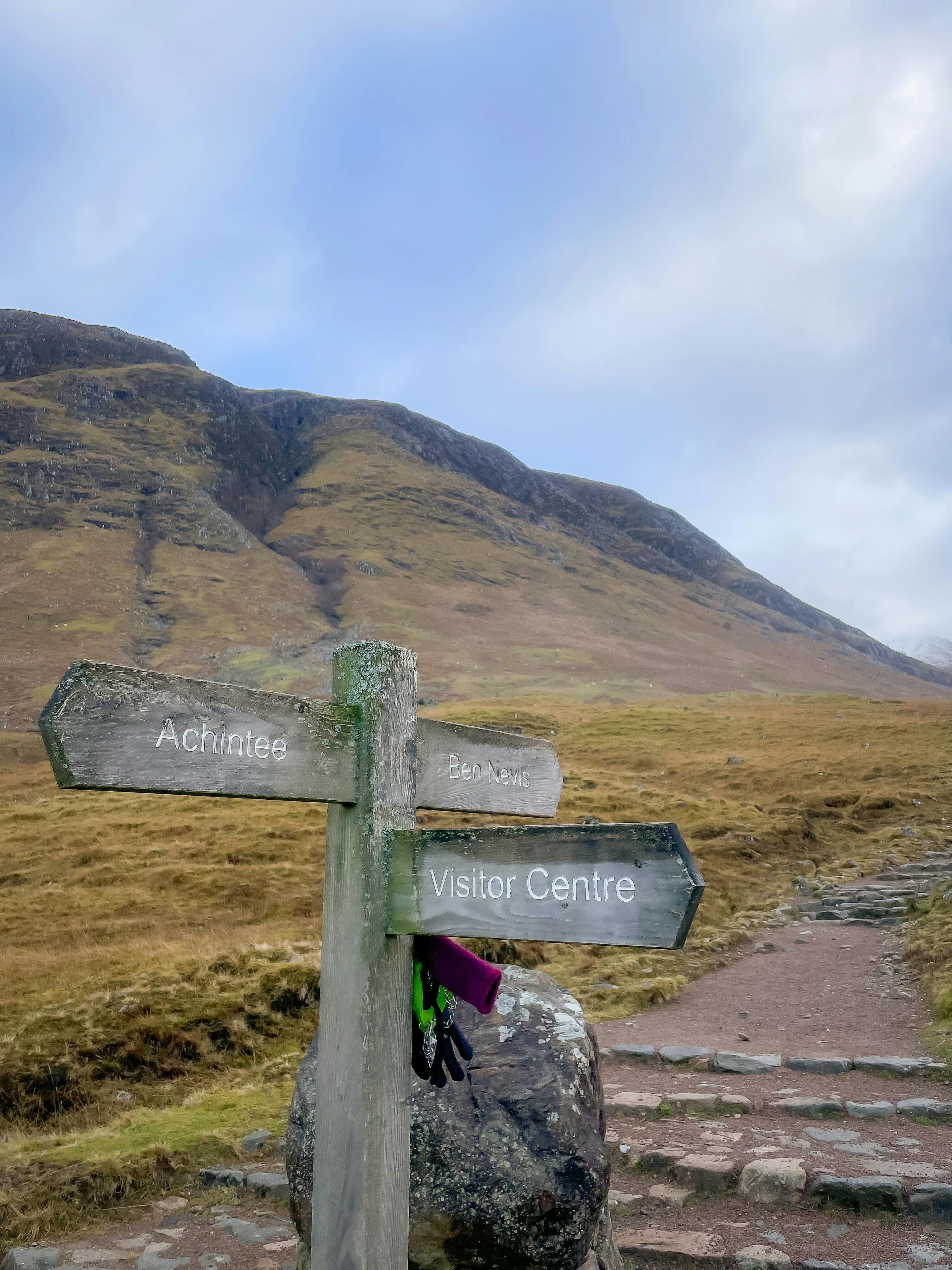A wooden signpost on a mountain trail pointing toward Achintee and the Visitor Centre.