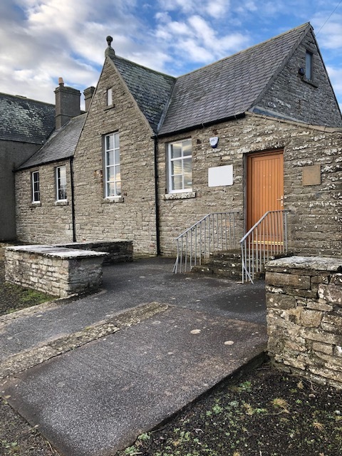 A stone building with a pitched roof and a wooden door, accessed by a small ramp and handrail.