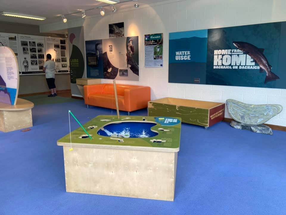 Interactive visitor centre room with wall displays about water and nature, orange seating, and a fishing game table.