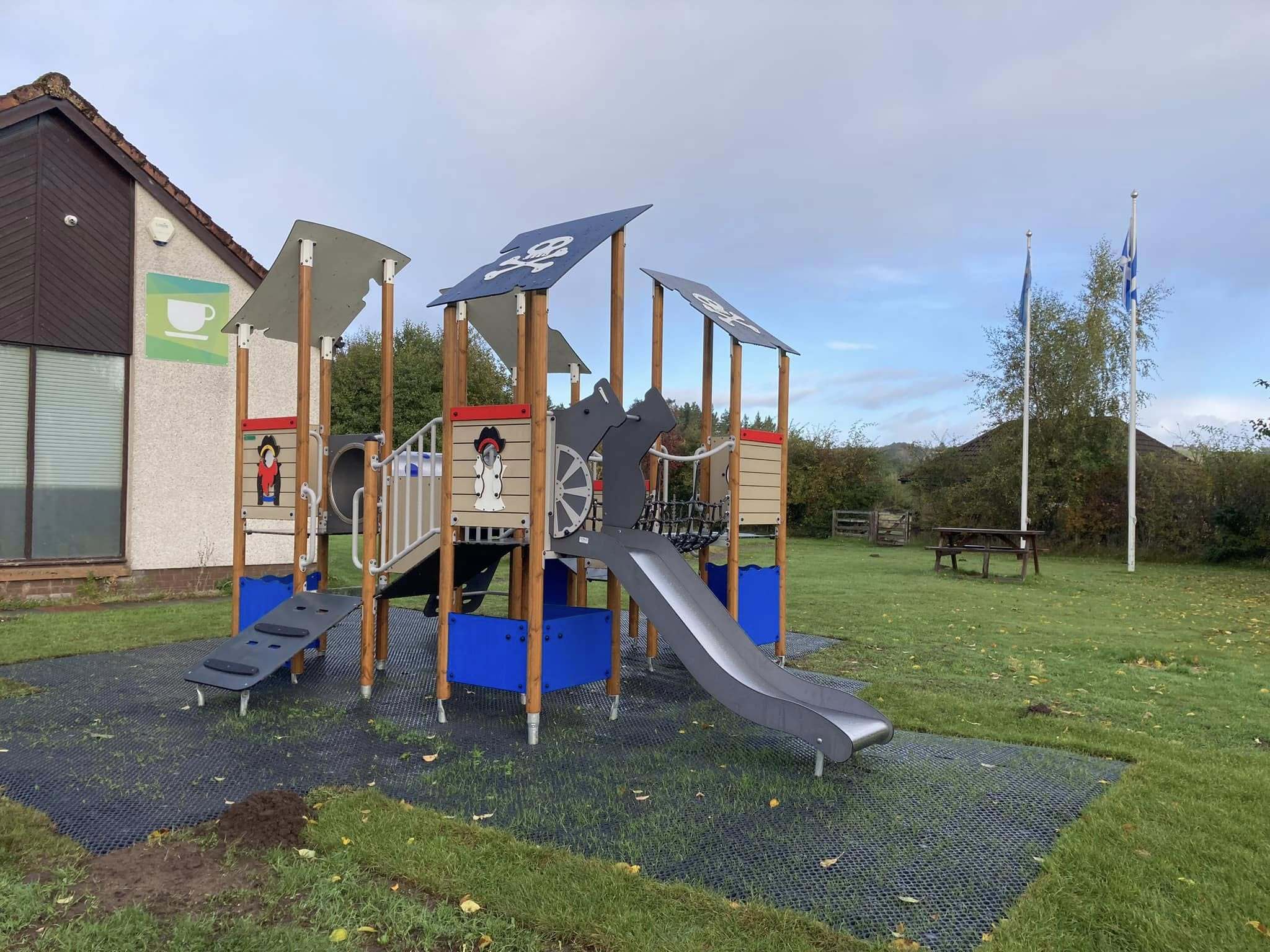 Outdoor pirate-themed play structure with slides, climbing platforms, and decorative sails, set on grass beside a building with a coffee sign and picnic benches in the background.