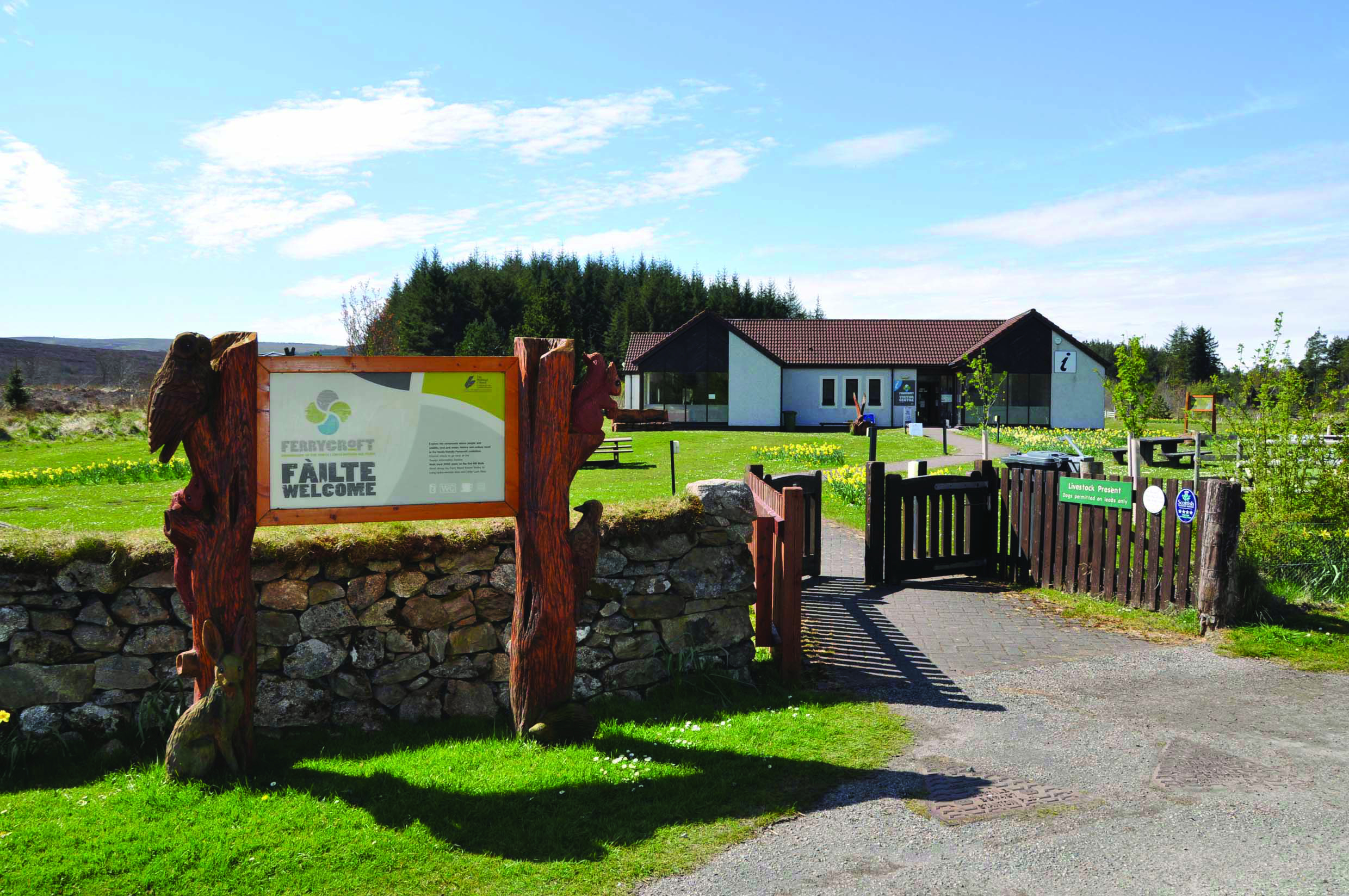 Entrance to Ferrycroft Visitor Centre on a sunny day, showing a stone wall with carved wooden posts framing a welcome sign, an open wooden gate, and a single-storey building with a red-tiled roof in the background, surrounded by green lawns and trees.