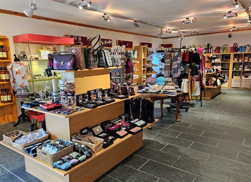Gift shop interior with shelves and tables displaying bags, scarves, accessories, and bottles of drinks.