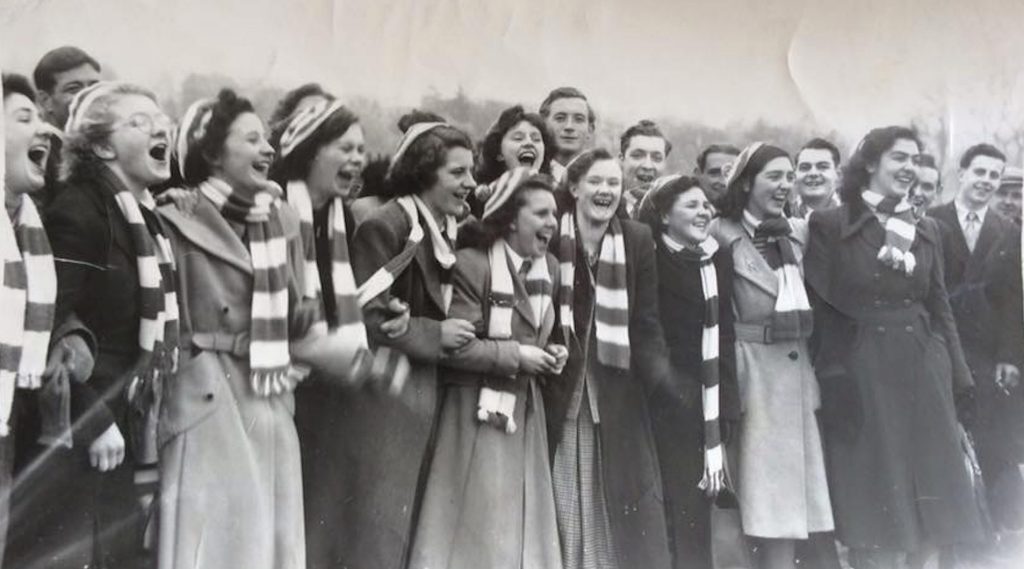 A group of women standing outdoors wearing striped scarves and coats, watching an event.