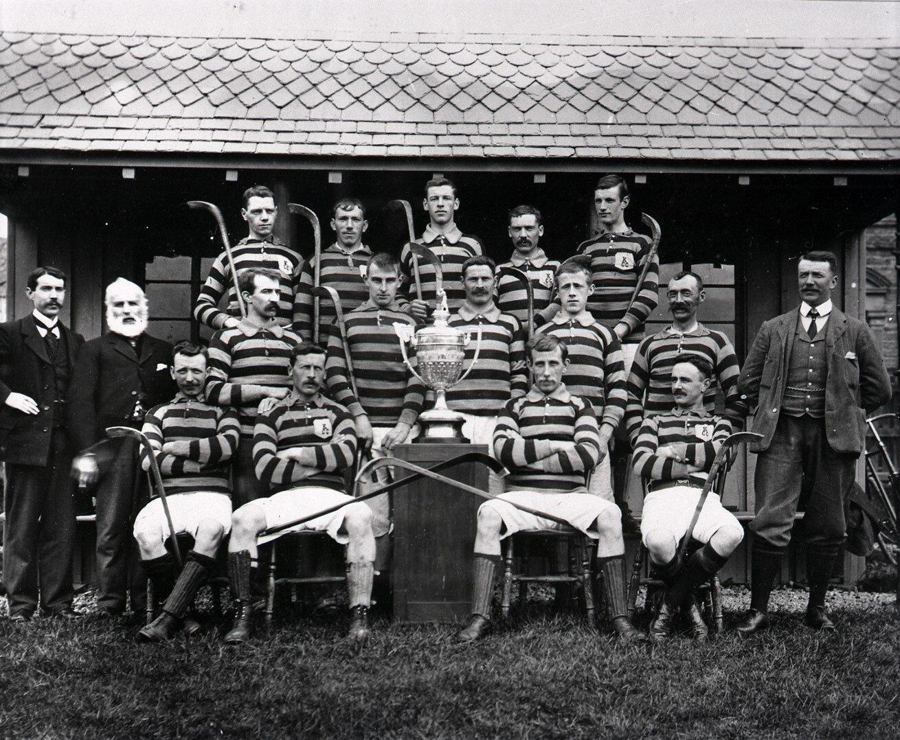 A shinty team posed in front of a pavilion, wearing striped jerseys and displaying a large trophy with camans arranged around it.