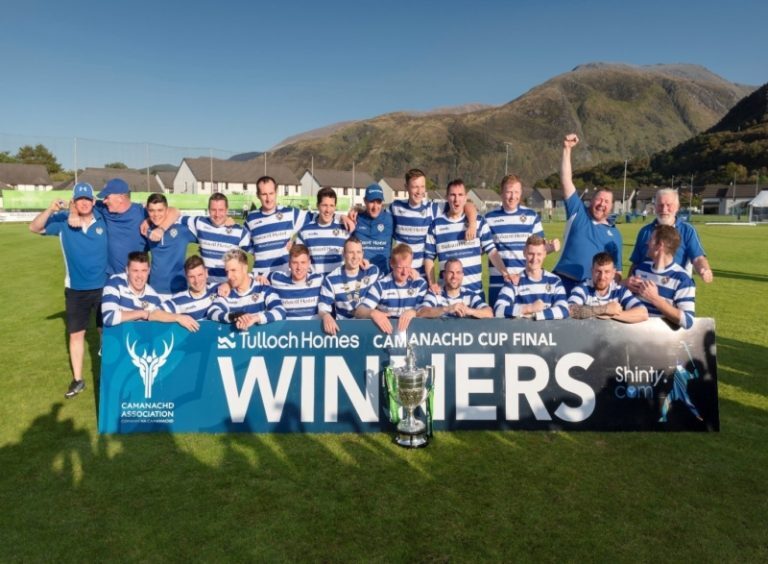 A shinty team in blue and white shirts posing behind a large “Winners” banner with the Camanachd Cup in front.
