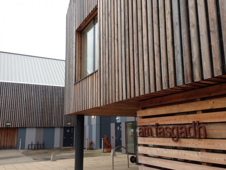 A close view of a wooden‑clad building with large windows and the sign “am fasgadh” beside the entrance.