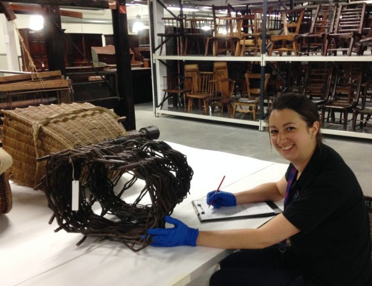 A person wearing blue gloves records notes on a clipboard while examining a large woven basket‑like object in a museum storage area filled with shelves of furniture.