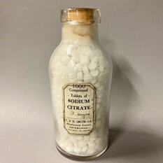 A small glass bottle filled with white tablets and sealed with a cork, labelled “Sodium Citrate.”