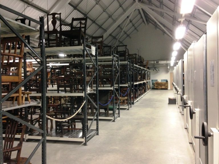 A long aisle of metal shelving filled with rows of wooden chairs inside a museum storage building.