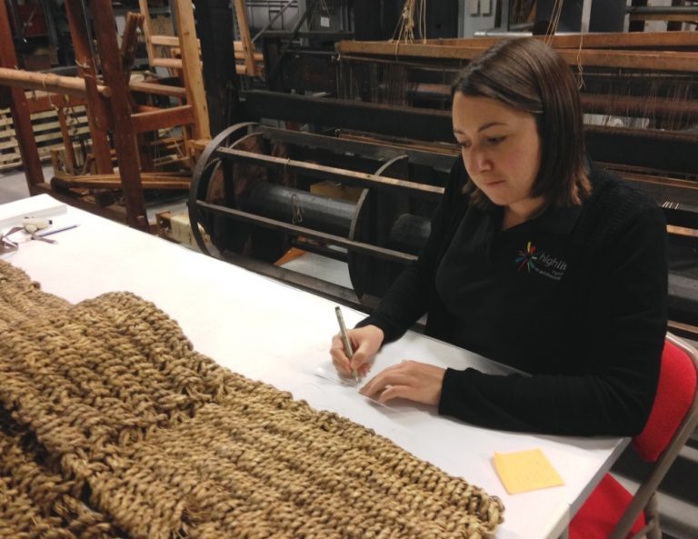 A person seated at a table writing notes beside a large thick woven mat in a museum store.