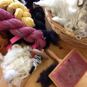 Skeins of dyed yarn, raw fleece, a drop spindle and a wool carder arranged on a table.