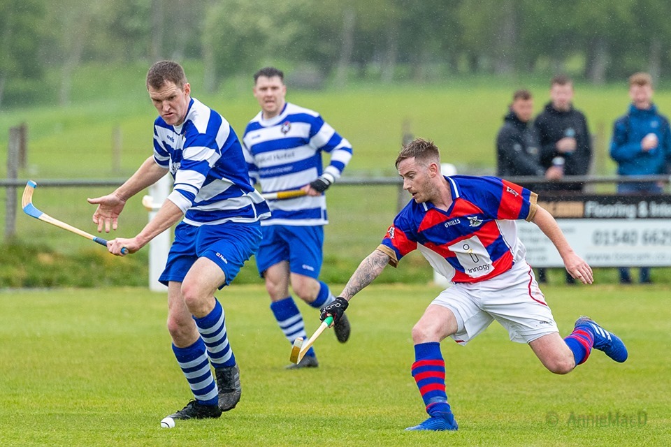Two shinty players competing for the ball during a match in the rain, one in blue and white stripes and the other in red and blue.